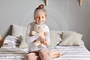 Happy smiling little girl with bun hairstyle wearing casual domestic clothing sitting on bed embracing her toy with love
