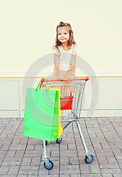 Happy smiling little girl child sitting in trolley cart with colorful shopping bags