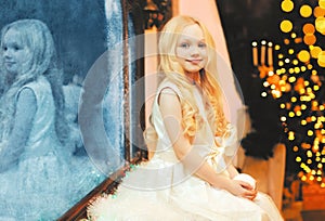 Happy smiling little girl child with snowball toy over christmas tree and garland lights at home near frozen mirror