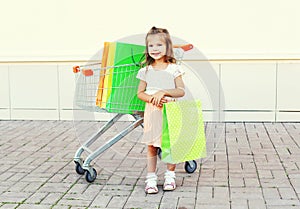 Happy smiling little girl child and trolley cart with shopping bags in city