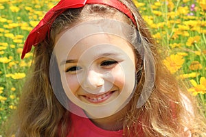 Happy smiling little girl with a red bow in her curly hair. Girl on a green meadow among yellow flowers