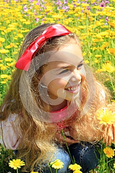 Happy smiling little girl with a red bow in her curly hair. Girl on a green meadow among yellow flowers