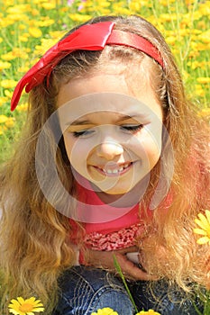 Happy smiling little girl with a red bow in her curly hair. Girl on a green meadow among yellow flowers