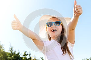 Happy smiling little girl in sunglasses, white t-shirt and summer hat against blue sky background