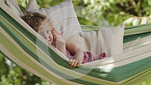 Happy smiling little girl waving hello, child with blue eyes and curly hair enjoys playing looking at camera. Lying on hammock in