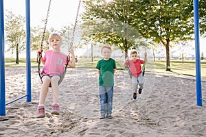 Happy smiling little preschool girl and boys friends swinging on swings at playground outside on summer day. Happy childhood