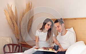 Happy and smiling in love young couple having romantic breakfast in bed on a sunday morning inside a house