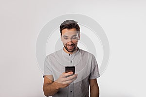 Happy smiling man in casual wear watching video on mobile phone while standing isolated in studio