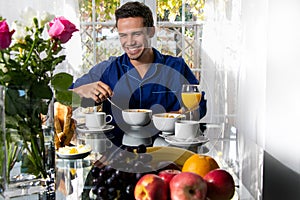 Happy smiling man in his pijamas having breakfast at table in front of patio doors with view of garden.