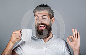 Happy smiling man holding cup of tasty fresh coffee and showing thumb up. Man holds cup of coffee, tea. Bearded man