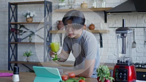 Happy smiling man holding veggie healthful smoothie in hand talking at tablet video chat. Portrait of cheerful Asian
