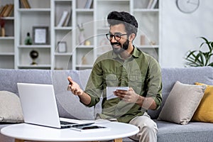 Happy and smiling man studying remotely sitting on sofa in living room, Hispanic man talking on video call using laptop