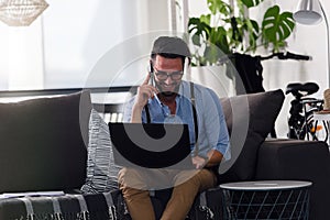 Happy smiling man using smartphone working on laptop computer while sitting on sofa