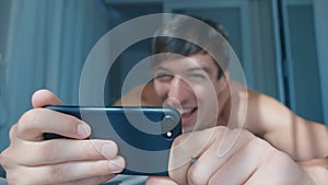 Happy smiling man watching video on smartphone lying in bed at home. Naked laughing male using smart phone playing game