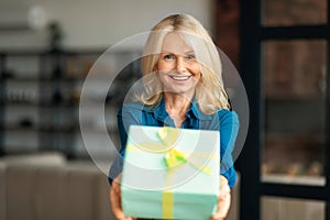 Happy smiling mature woman giving wrapped box with present, stretching hands to camera, home interior