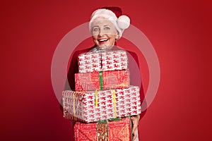 mature woman in red santa claus hat holding stack of gift boxes, on red studio background. Christmas shopping, copy