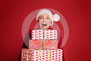 Mature woman in red santa claus hat holding stack of gift boxes, on red studio background. Christmas shopping, copy