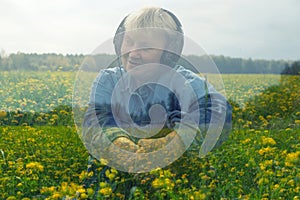 Happy smiling mature woman sitting wearing headphones and listen to relaxation music on field with alot of yellow flowers. Double