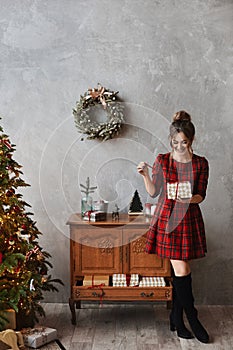 Happy smiling model girl in plaid red dress holding the present box and posing near Christmas tree. Young beautiful