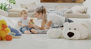 Happy and smiling mom with child baby playing with wooden toys and plush stuffed animals, at home sitting on floor with pillows,