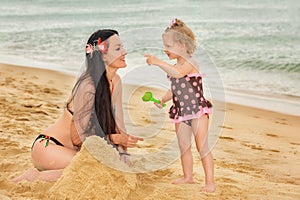 Happy smiling mom with a daughter child play on the beach and build sand castles on a sunny summer day. Mother and girl