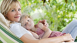 Happy and smiling mom hugs her little girl daughter child blue eyes with blond curly hair, together lying on the hammock in the