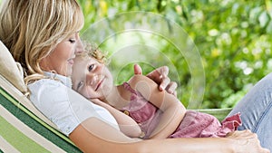 Happy and smiling mom hugs her little girl daughter child blue eyes with blond curly hair, together lying on the hammock in the