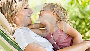 Happy and smiling mom hugs her little girl daughter child blue eyes with blond curly hair, together lying on the hammock in the