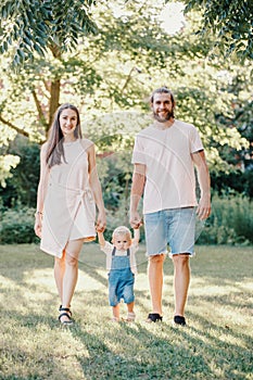 Happy smiling mother and father hold hands with baby boy in park outdoor. Family Caucasian mom and dad with son walking together.