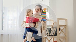 Happy smiling mother giving toys to her baby son from the shelf in living room. Baby development, family playing games, making