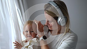 Happy smiling mother in headphones looking out the window caressing head of joyful baby girl posing laughing. Loving