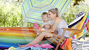 Happy smiling mother with her little baby son, together they look at mobile phone and take a selfie on a sunny summer day, mom