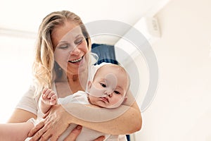 Happy smiling mother holds and hugs her baby in a bright room. Harmonious family, happy motherhood and childhood.