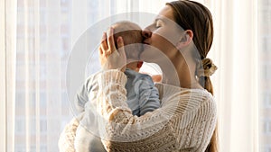 Happy smiling mother hugging and kissing her little baby son at big window in living room. Concept of family happiness