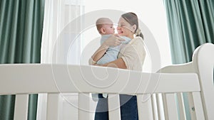 Happy smiling mother hugging and kissing her little baby son at morning standing in bedroom with big window. Concept of