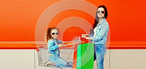 Happy smiling mother and little girl child with trolley cart and shopping bags in the city
