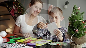 Happy smiling mother looking at her son making traditional Christmas postcard and handmade decorations