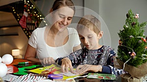 Happy smiling mother looking at her son making traditional Christmas postcard and handmade decorations