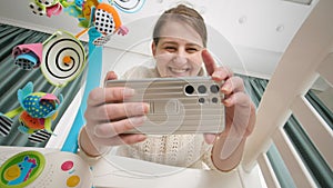Happy smiling mother making video on smartphone of her baby lying in cradle. Concept of parenting and making images of