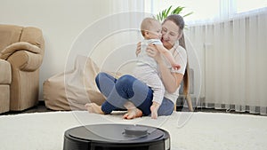 Happy smiling mother playing with her baby son while robot vacuum cleaner clean house. Concept of hygiene, household