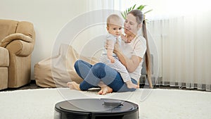 Happy smiling mother playing with her baby son while robot vacuum cleaner clean house. Concept of hygiene, household