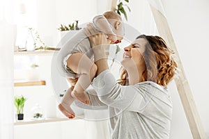 Happy smiling mother playing with newborn child in comfy light bedroom in front of window. Moments of motherhood