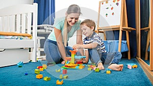 Happy smiling mother sitting on floor with her little son and playing with colorful constructro blocks