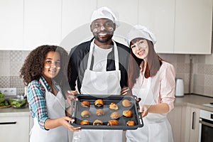 Happy smiling parents, African man, Caucasian woman and their mixed race teen child, holding baking tray with fresh