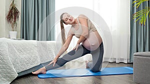 Happy smiling pregnant woman doing sportrs stretching and fitness exercises on mat at big window in morning. Concept of