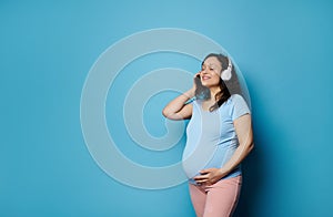 Happy smiling pregnant woman enjoying listening to soundtrack on white headphones, stroking her belly, isolated on blue