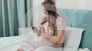 Happy smiling pregnant woman listening to music with headphones and using smartphone while sitting on bed at home