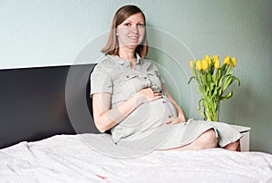 Happy smiling pregnant woman sitting on a bed in a bedroom at home and touching her belly.