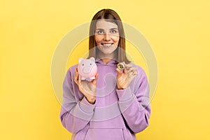 Happy smiling pretty woman with hair holding piggy bank and gold crypto coin, growth bitcoin.