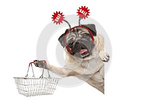 Happy smiling pug puppy dog, holding up shopping basket, wearing diadem with red sale sign